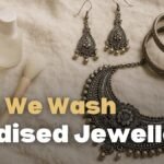 can we wash oxidised jewellery, A soft, crinkled off-white linen background with oxidized silver jewelry arranged on the right, including a floral statement necklace, dangling earrings, and chunky bangles. On the left, gentle cleaning tools are displayed: a ceramic bowl of clear water, a subtle bottle, a rolled microfiber cloth, and a small, soft-bristled jewelry cleaning brush with a wooden handle. The scene is lit with warm natural light, evoking a cozy, vintage boho aesthetic
