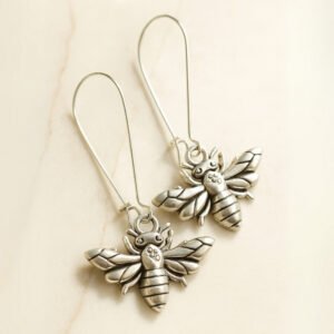 A pair of silver bee-shaped dangle earrings with long hooks displayed on a smooth cream background, highlighting the intricate detailing of the wings and body.